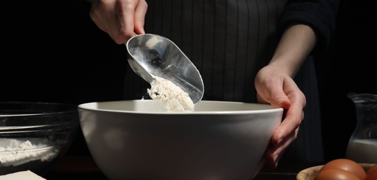 Photo of Making bread. Woman adding flour into bowl at wooden table on dark background, closeup