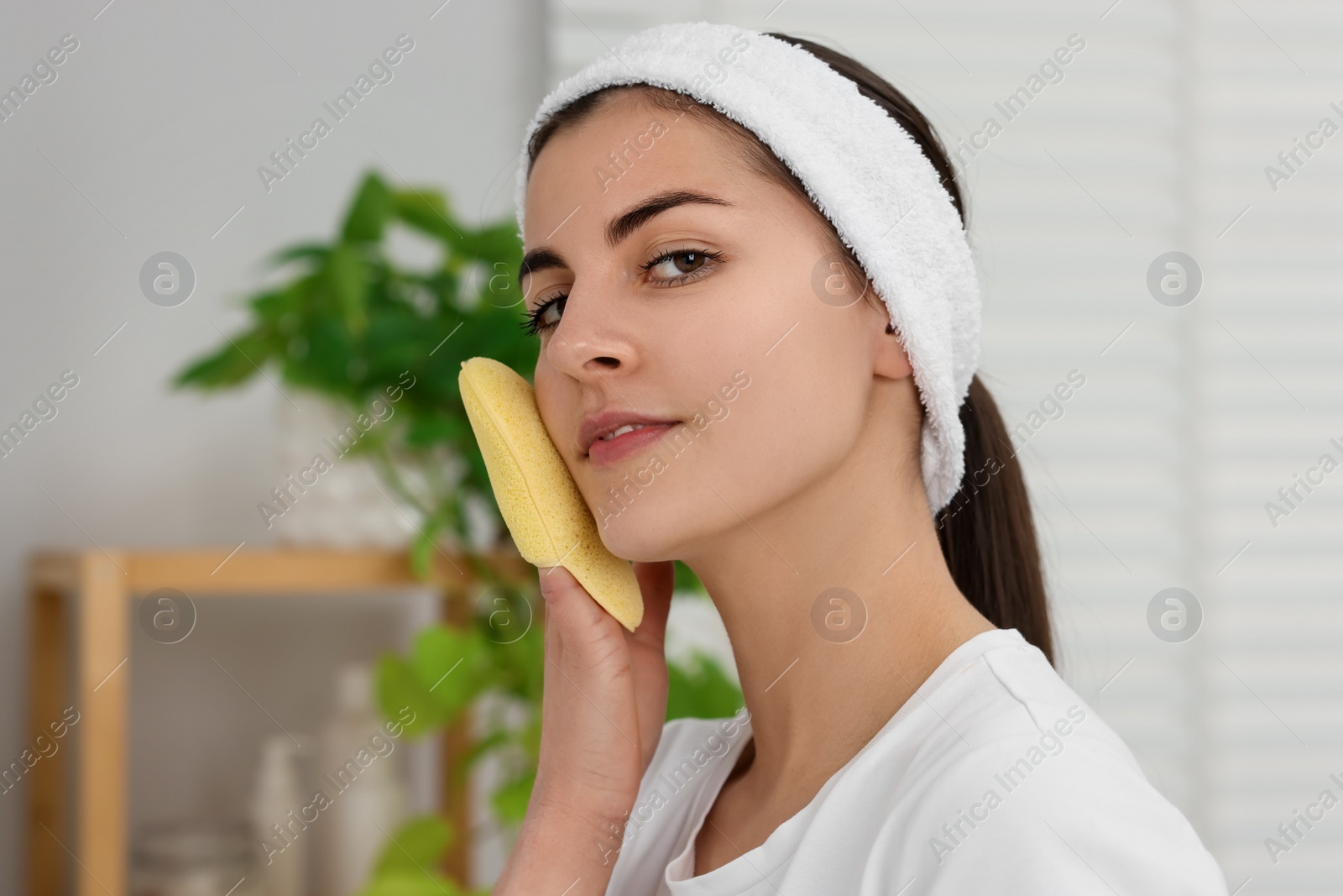 Photo of Young woman with headband washing her face using sponge in bathroom