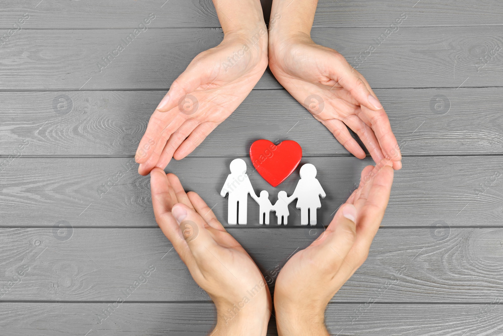 Photo of Young man and woman protecting family figure and red heart with their hands on grey wooden background, top view