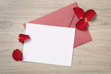 Photo of Blank greeting card, envelope and rose petals on wooden table, flat lay. Valentine's day celebration