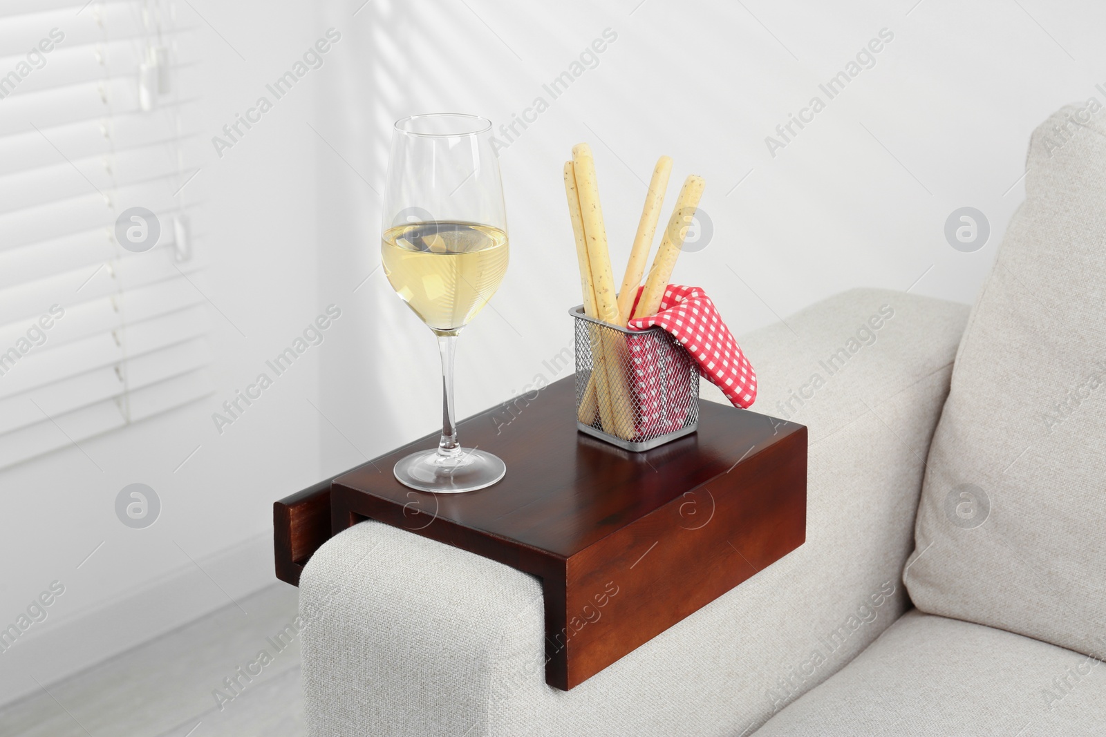 Photo of Glass of white wine and breadsticks on sofa with wooden armrest table in room. Interior element