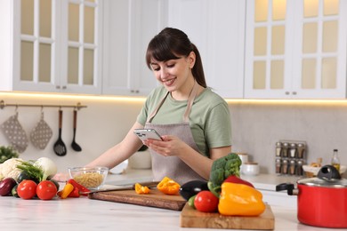 Happy young housewife using smartphone while cooking at white marble table in kitchen