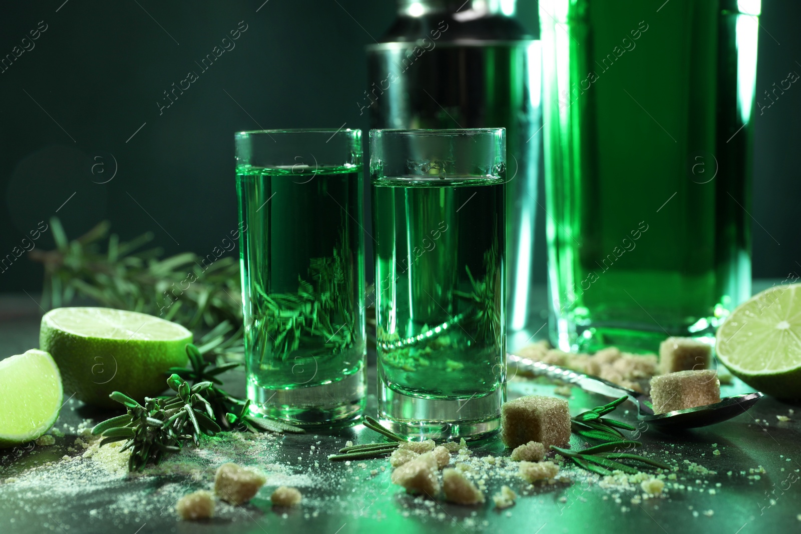 Photo of Absinthe in shot glasses, spoon, brown sugar, lime and rosemary on gray table, closeup. Alcoholic drink