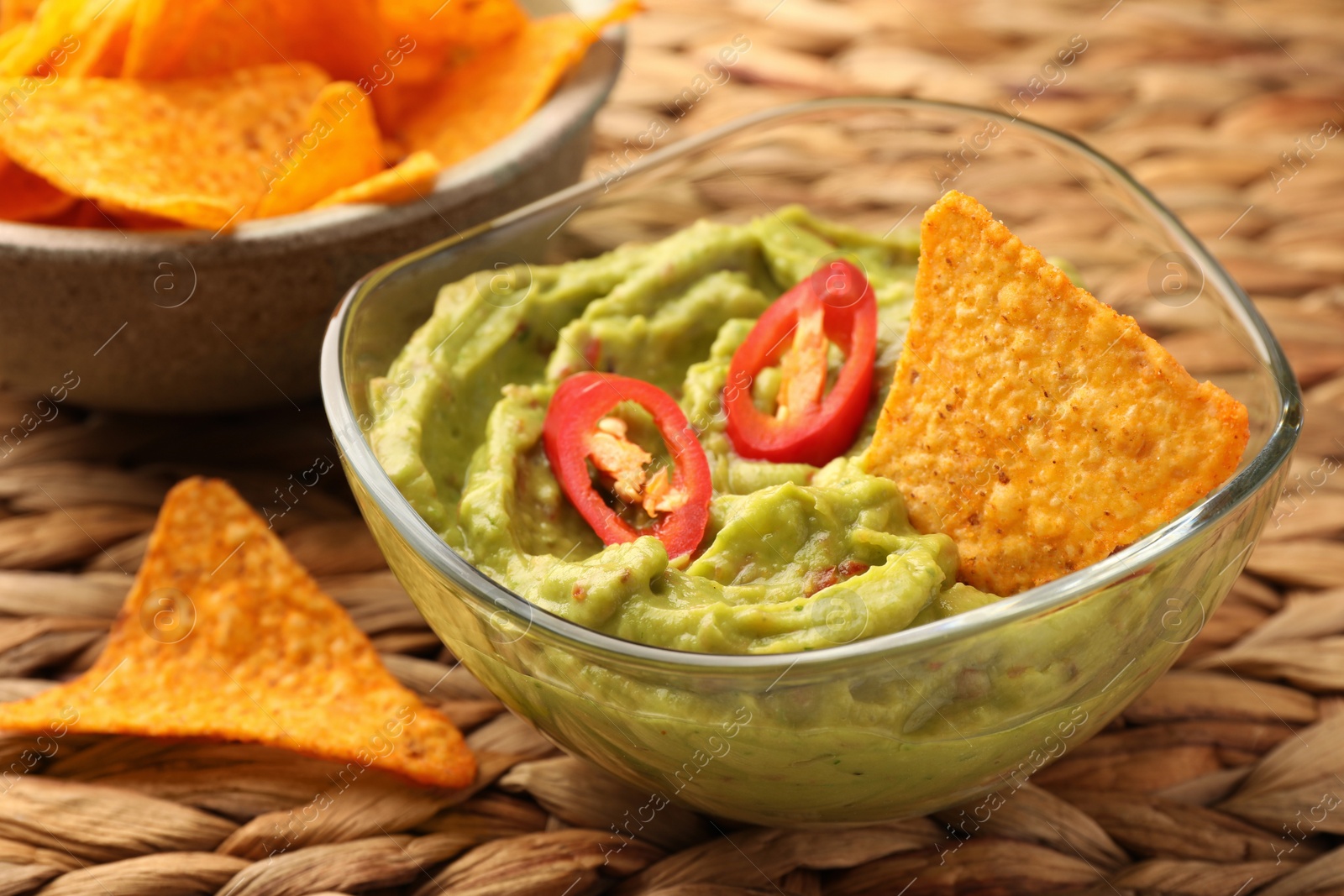Photo of Bowl of delicious guacamole with chili pepper and nachos chips on wicker table, closeup