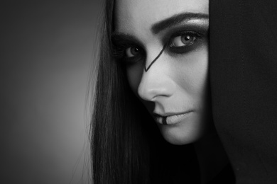 Mysterious witch in mantle with hood on dark background, closeup. Black and white effect