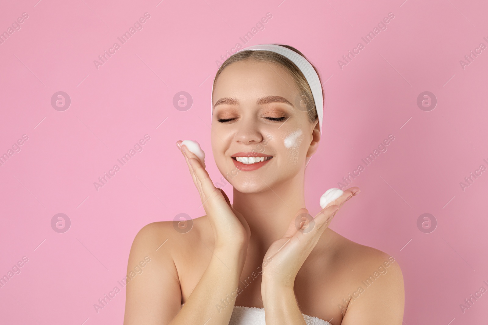 Photo of Young woman washing face with cleansing foam on pink background. Cosmetic product