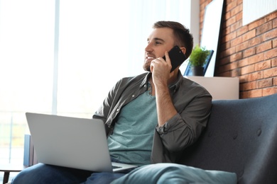 Photo of Young man talking on phone while using laptop in living room