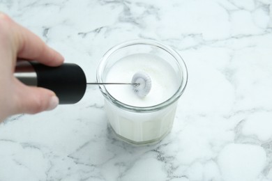 Photo of Woman whisking milk in glass with mini mixer (milk frother) at white marble table, closeup