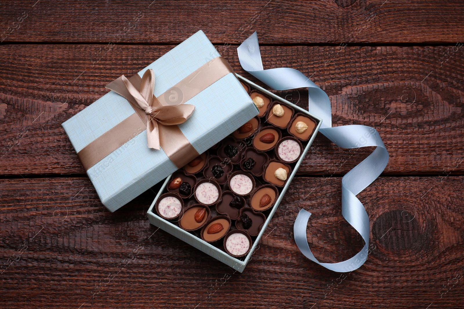 Photo of Open box of delicious chocolate candies and light blue ribbon on wooden table, flat lay
