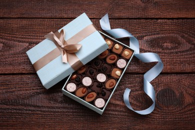 Open box of delicious chocolate candies and light blue ribbon on wooden table, flat lay