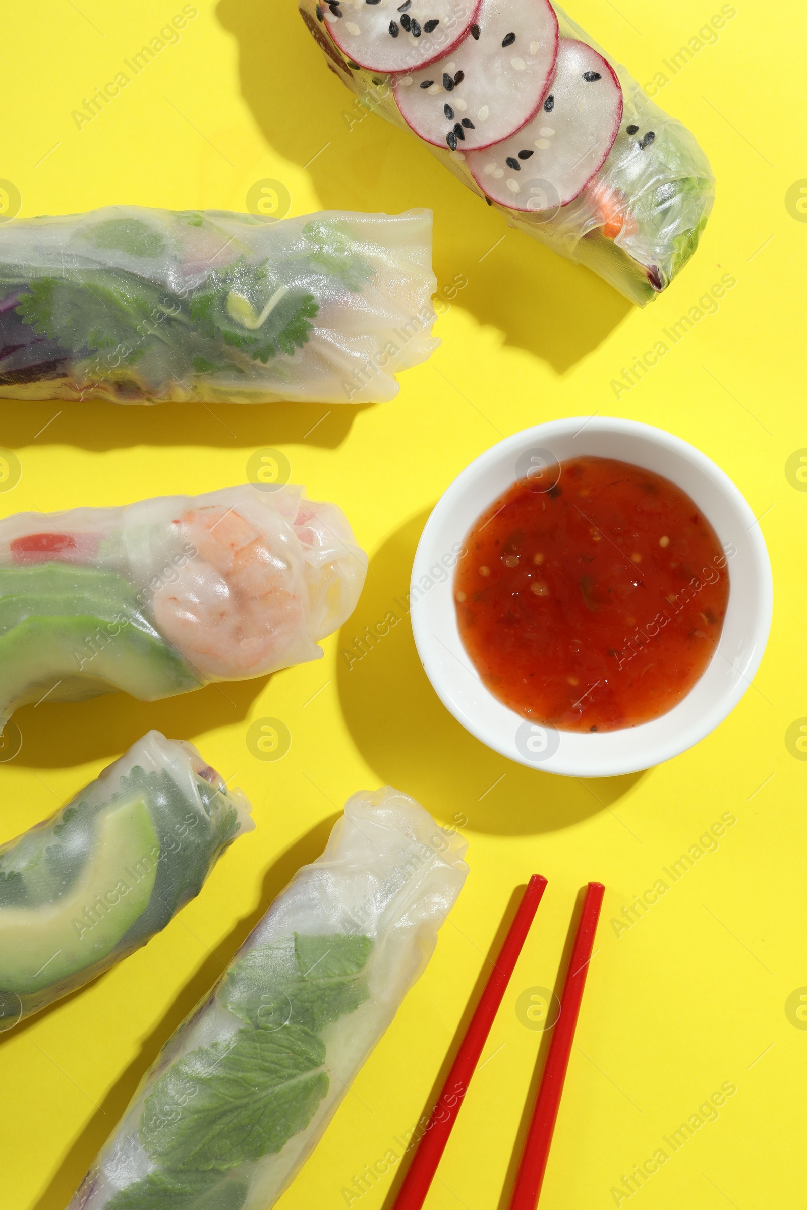 Photo of Delicious spring rolls, sauce and chopsticks on yellow background, flat lay