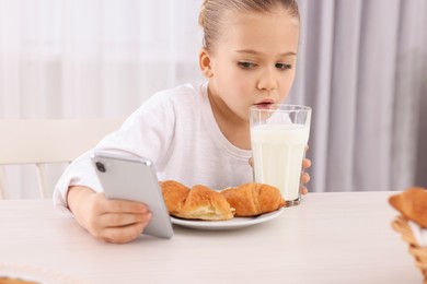Little girl using smartphone while having breakfast at table indoors. Internet addiction