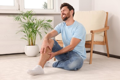 Man suffering from leg pain and touching knee near soft armchair at home