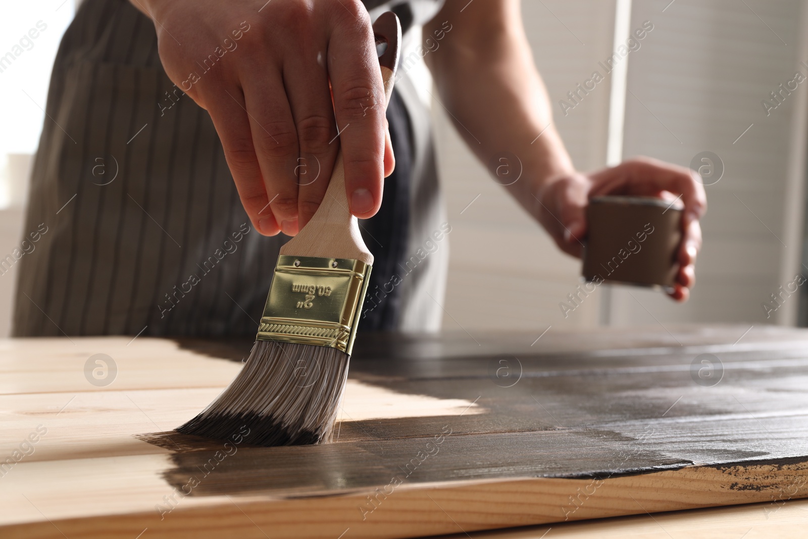 Photo of Man with brush and can applying wood stain onto wooden surface indoors, closeup