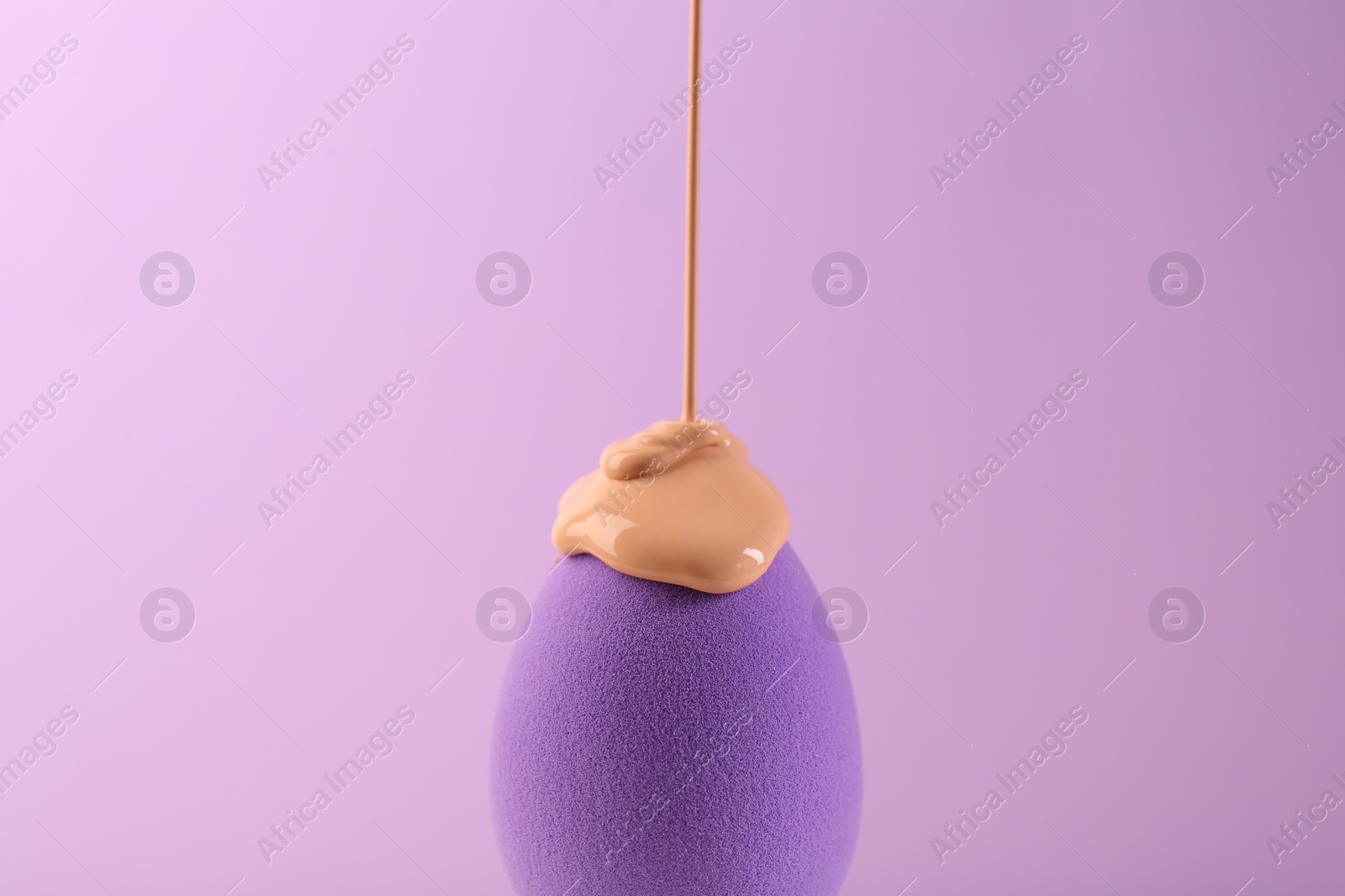 Photo of Pouring skin foundation onto makeup sponge on violet background, closeup