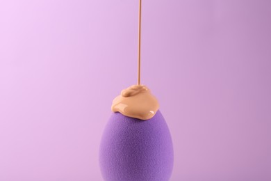 Pouring skin foundation onto makeup sponge on violet background, closeup