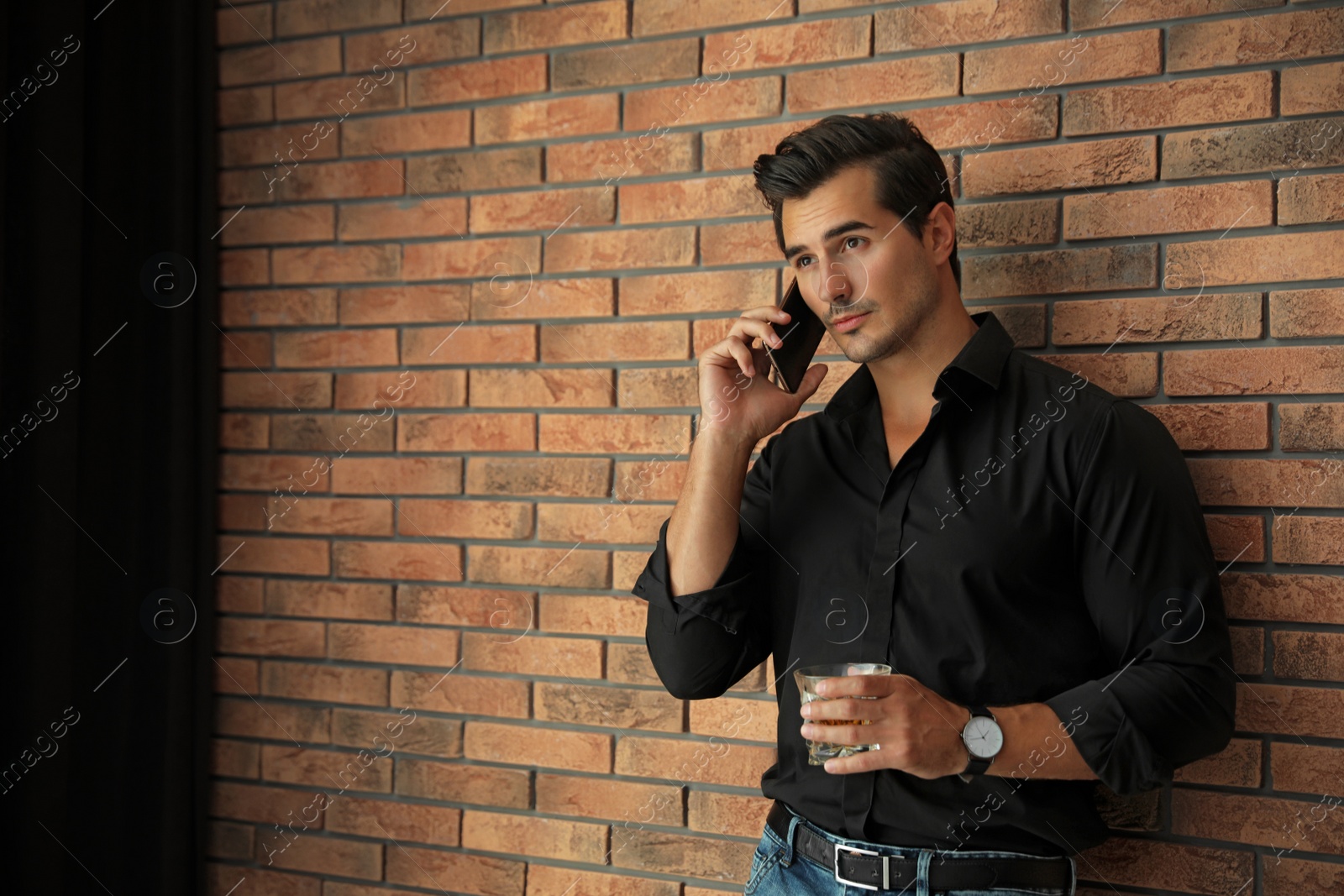 Photo of Young man with glass of whiskey talking on phone near brick wall indoors. Space for text