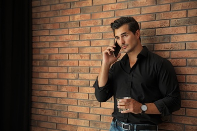 Young man with glass of whiskey talking on phone near brick wall indoors. Space for text