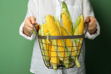 Photo of Woman holding metal basket with tasty fresh corn cobs on green background, closeup