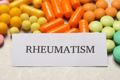 Photo of Paper with word Rheumatism and pills on light marble table, closeup