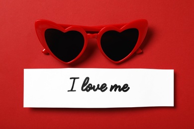 Photo of Paper with handwritten phrase I Love Me and heart shaped sunglasses on red background, flat lay