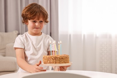 Cute boy with birthday cake at table indoors, space for text