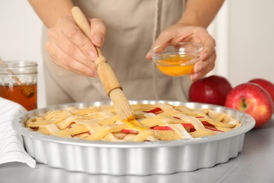 Woman applying liquid egg onto traditional English apple pie with brush at light grey table, closeup