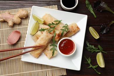 Photo of Tasty fried spring rolls, arugula, lime and sauces served on wooden table, flat lay