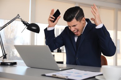 Emotional businessman with laptop and smartphone at table in office