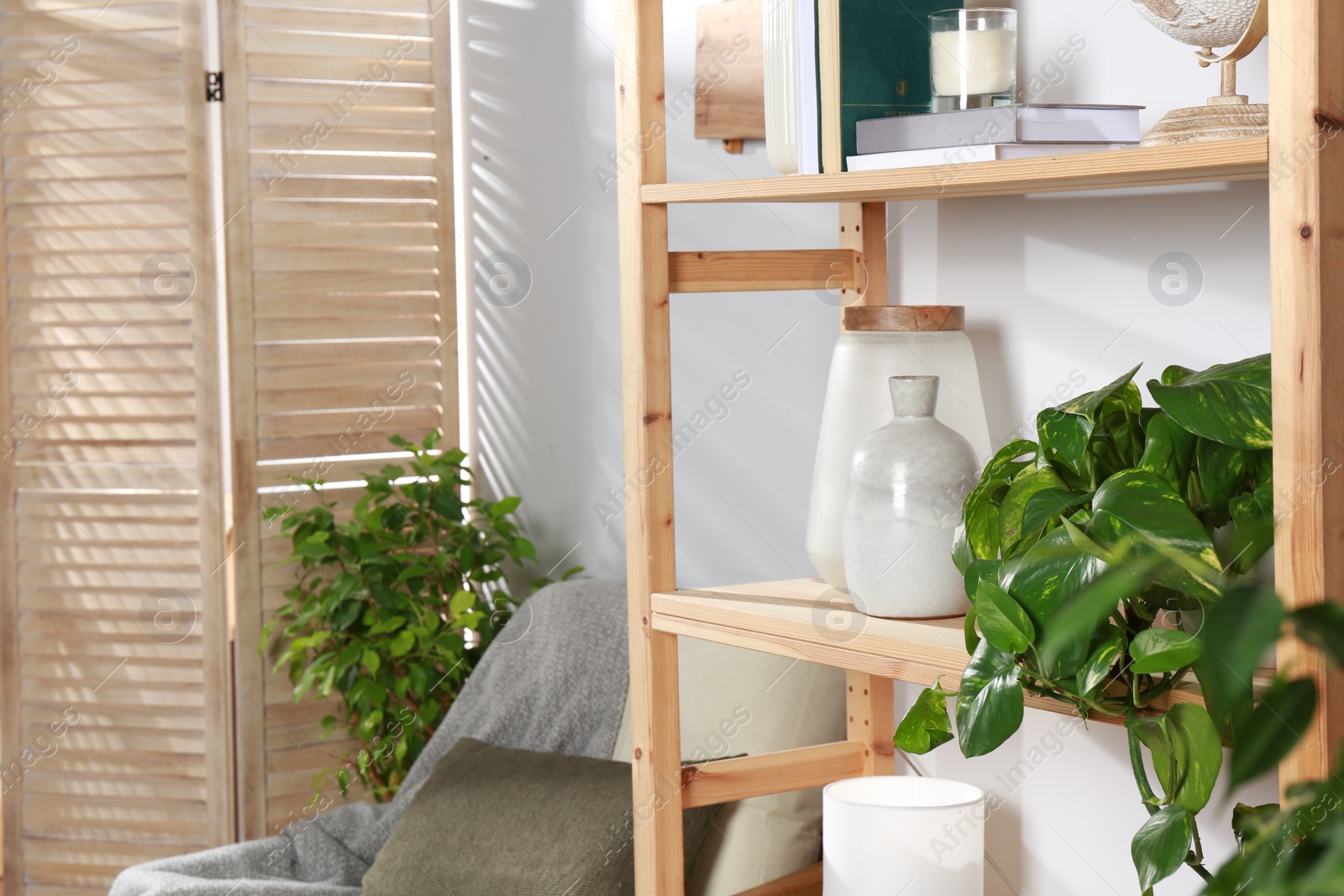 Photo of Wooden shelving unit with home decor, folding screen and armchair near light wall in room. Interior design