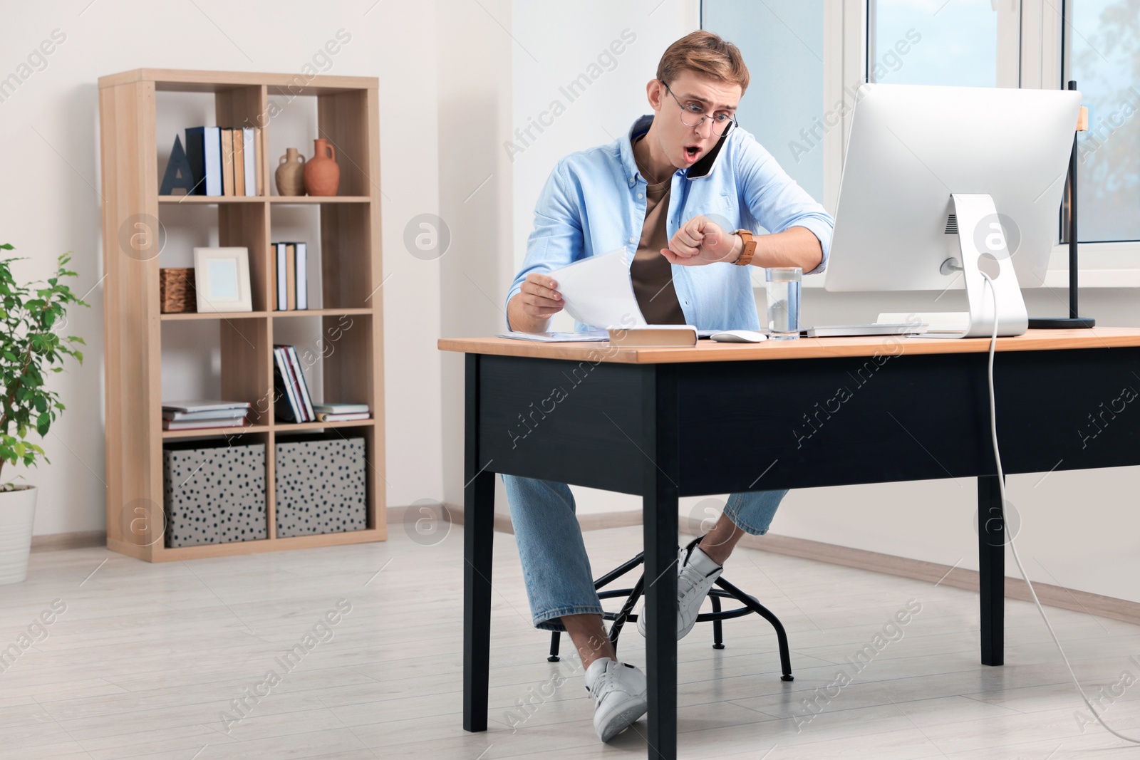 Photo of Emotional young man checking time while talking on phone in office. Being late