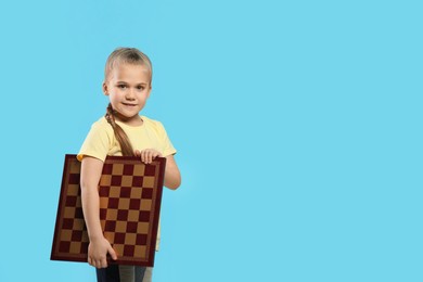 Photo of Cute girl holding chessboard on light blue background. Space for text