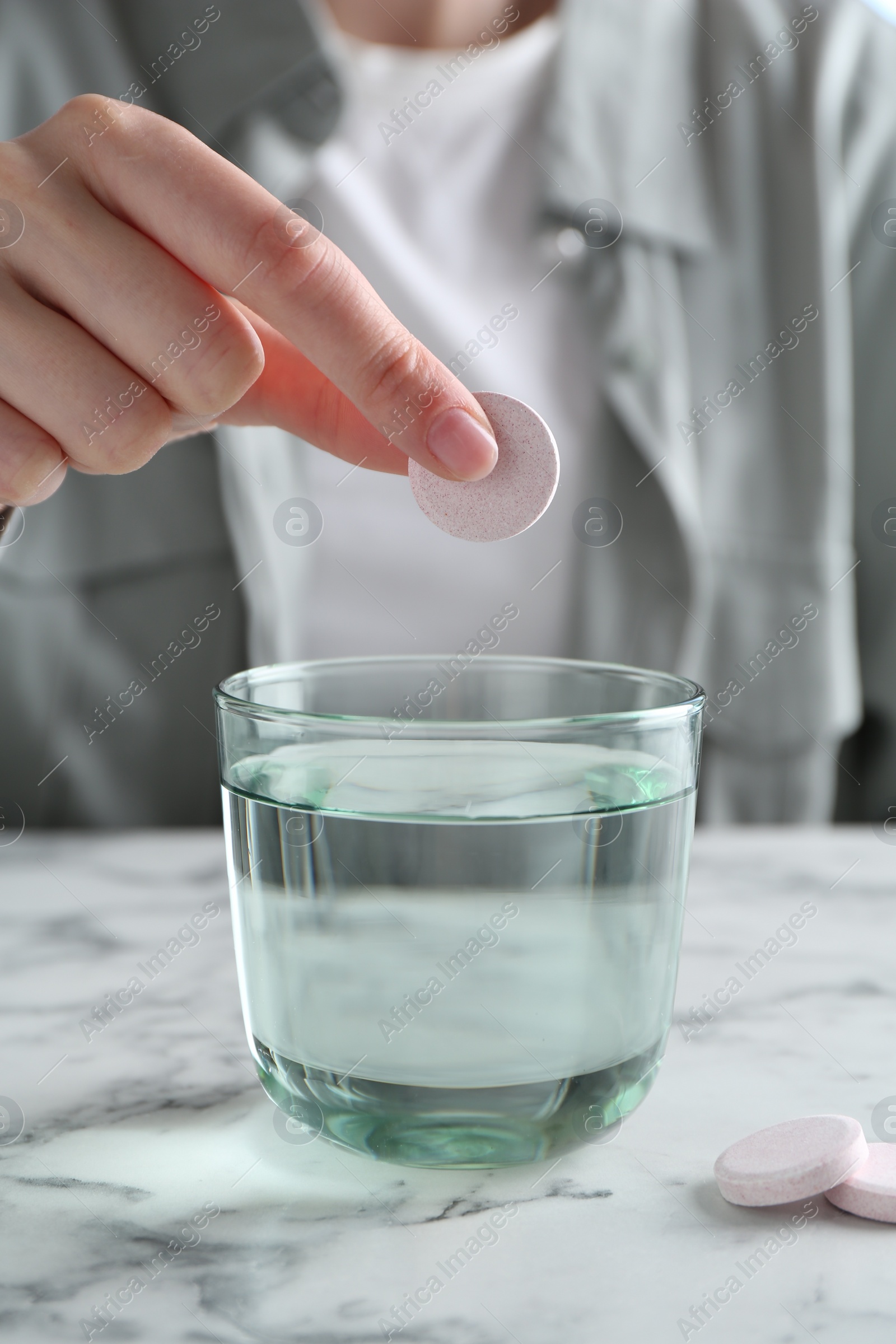 Photo of Woman putting effervescent pill into glass of water at white marble table, closeup