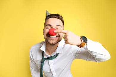Photo of Emotional young man with party cap and clown nose on yellow background. April fool's day