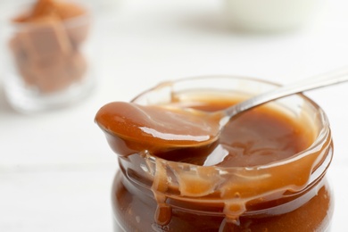 Jar and spoon with caramel sauce on blurred background, closeup