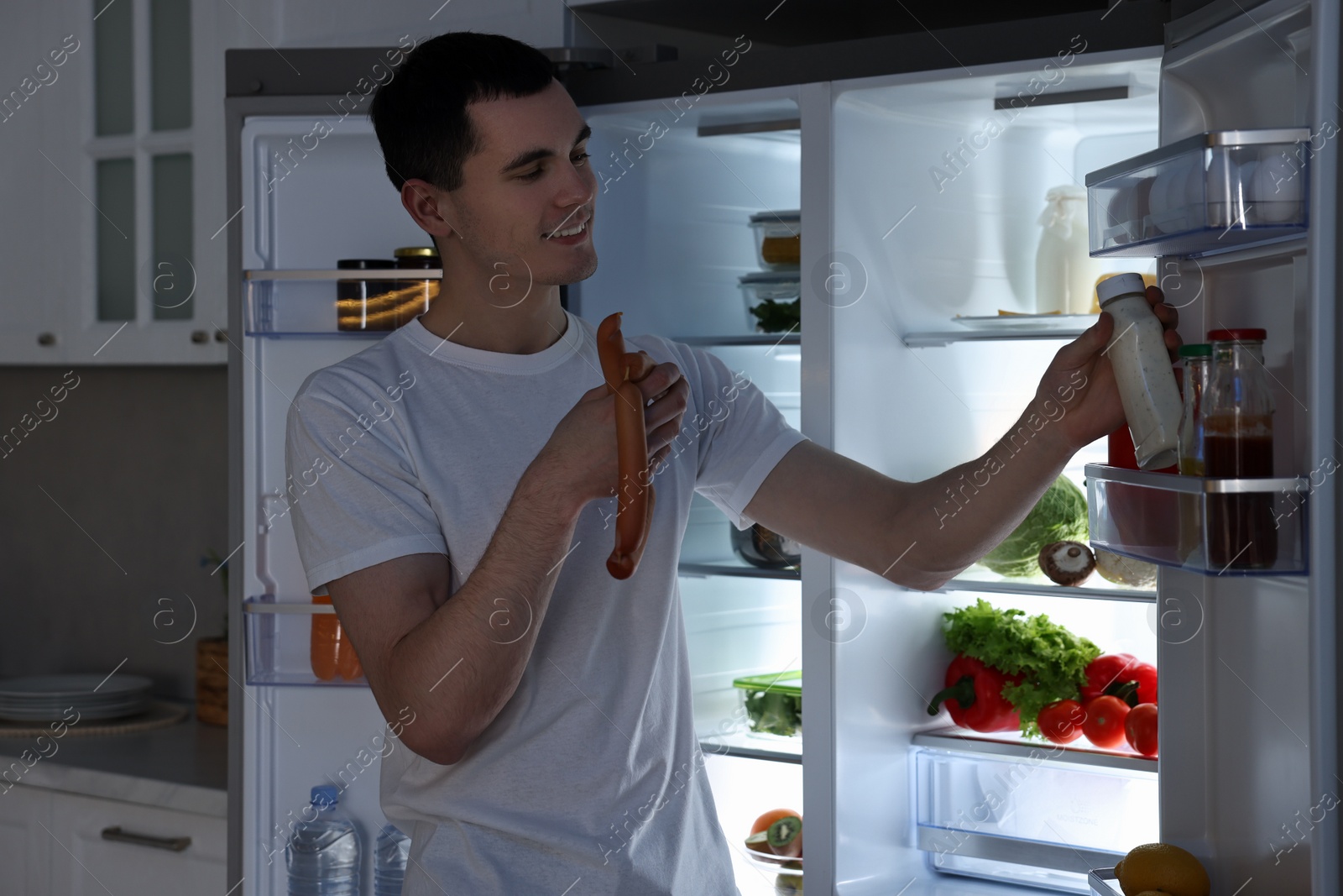 Photo of Happy man with sausages taking sauce out of refrigerator in kitchen at night