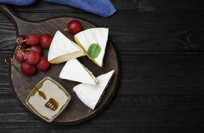 Tasty brie cheese with basil, grapes and honey on black wooden table, flat lay. Space for text