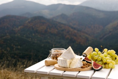 Photo of Delicious cheese, nuts and fruits on white wooden table against mountain landscape. Space for text