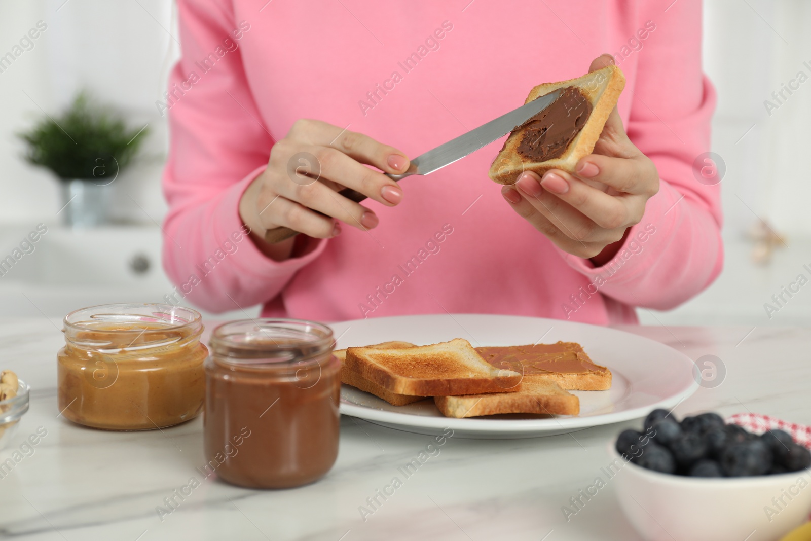 Photo of Woman spreading tasty nut butter onto toast at white marble table, closeup