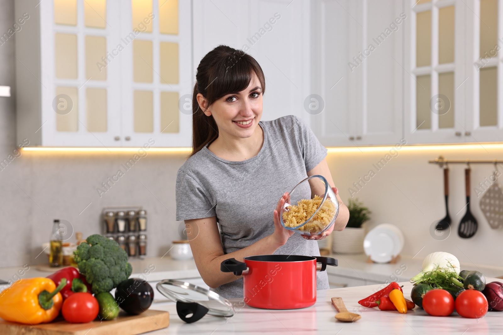 Photo of Happy young housewife adding raw pasta in pot at white marble table in kitchen. Cooking process