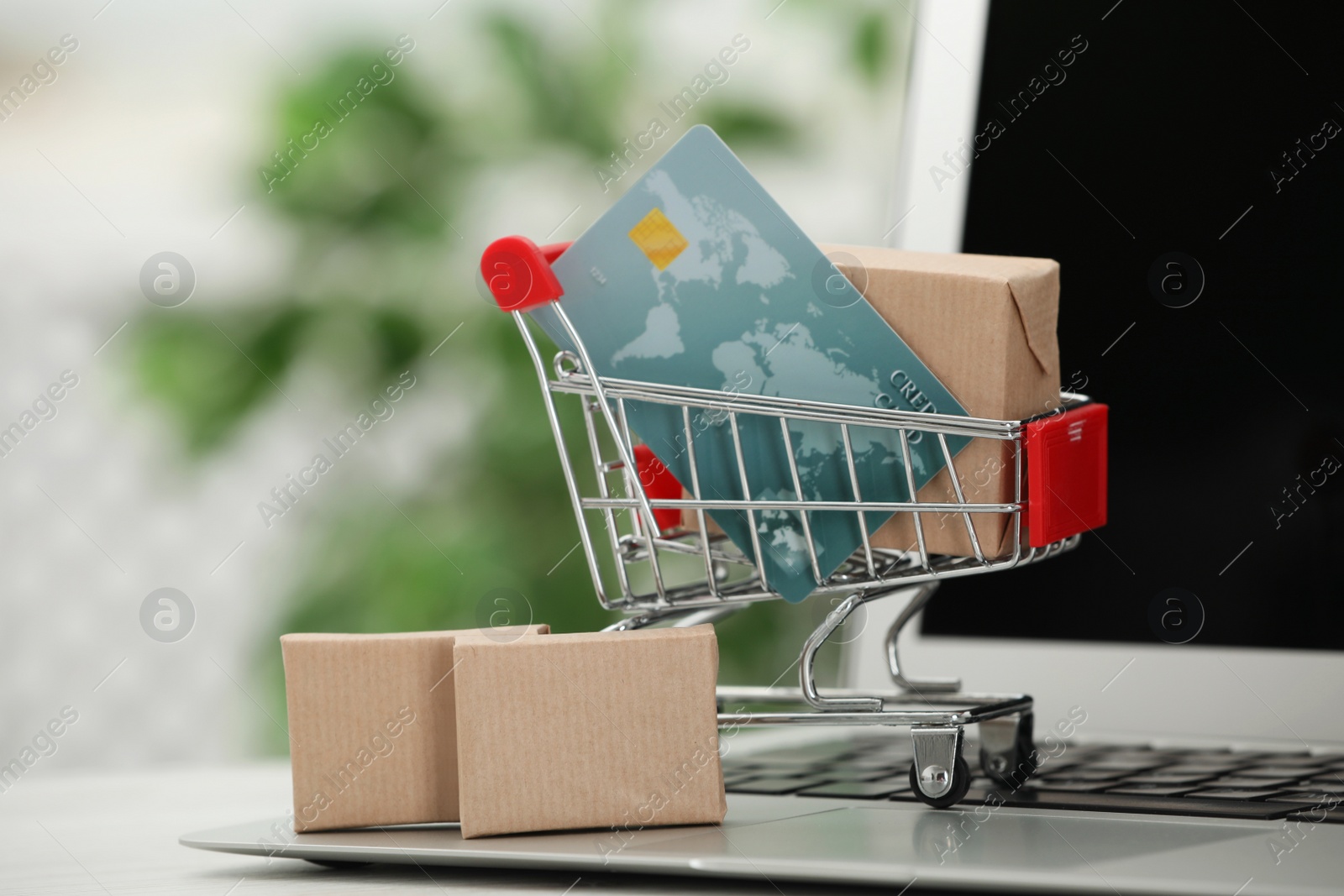 Photo of Online payment concept. Small shopping cart with bank card, boxes and laptop on table, closeup