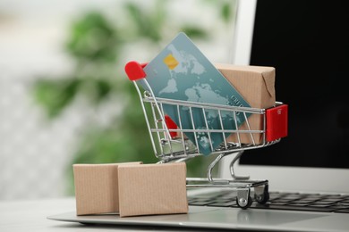 Photo of Online payment concept. Small shopping cart with bank card, boxes and laptop on table, closeup