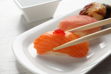 Taking delicious nigiri sushi with chopsticks from plate on white wooden table, closeup