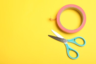 Photo of Roll of pink adhesive tape and scissors on yellow background, flat lay. Space for text