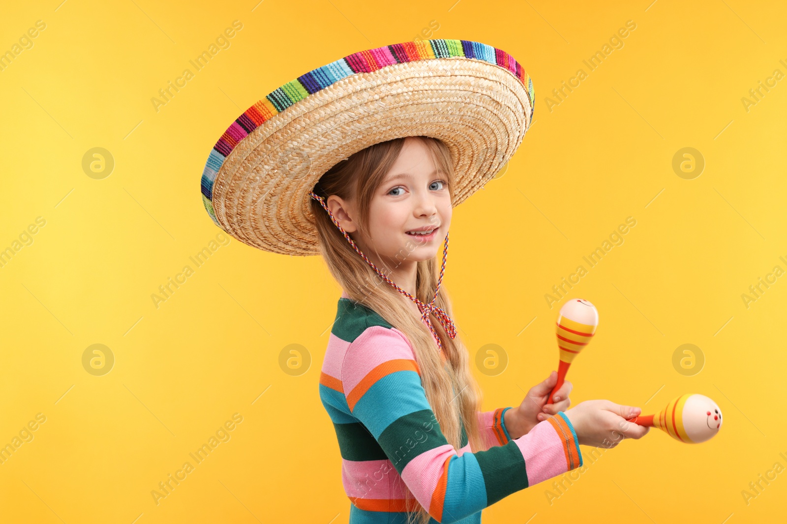 Photo of Cute girl in Mexican sombrero hat dancing with maracas on orange background