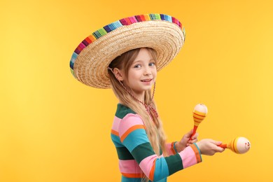 Cute girl in Mexican sombrero hat dancing with maracas on orange background