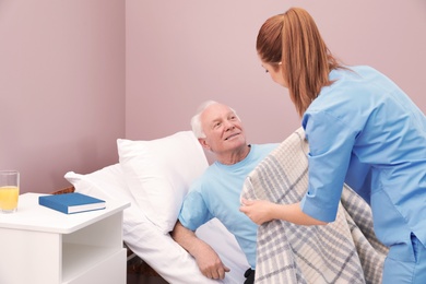 Photo of Nurse covering senior man with plaid on bed in hospital ward. Medical assisting
