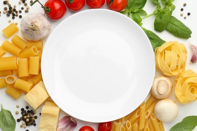 Photo of Plate surrounded by different types of pasta, products and peppercorns on white background, flat lay. Space for text
