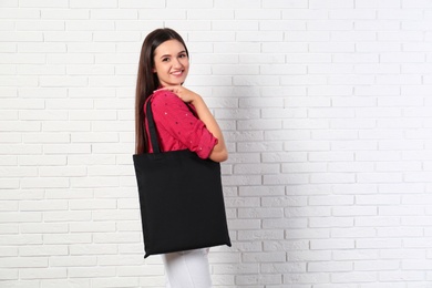 Photo of Beautiful young woman with stylish blank eco bag near brick wall, space for text