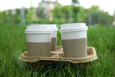 Photo of Takeaway paper coffee cups with plastic lids and sleeves in cardboard holder on green grass outdoors
